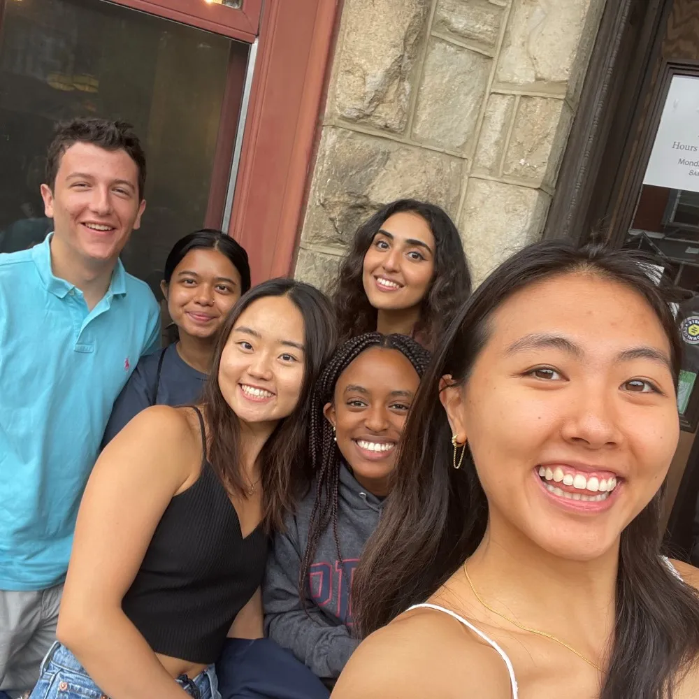 Six young people smiling together for a group selfie outside a building.